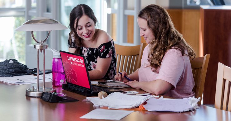 two female students at a desk with a computer and a laptop