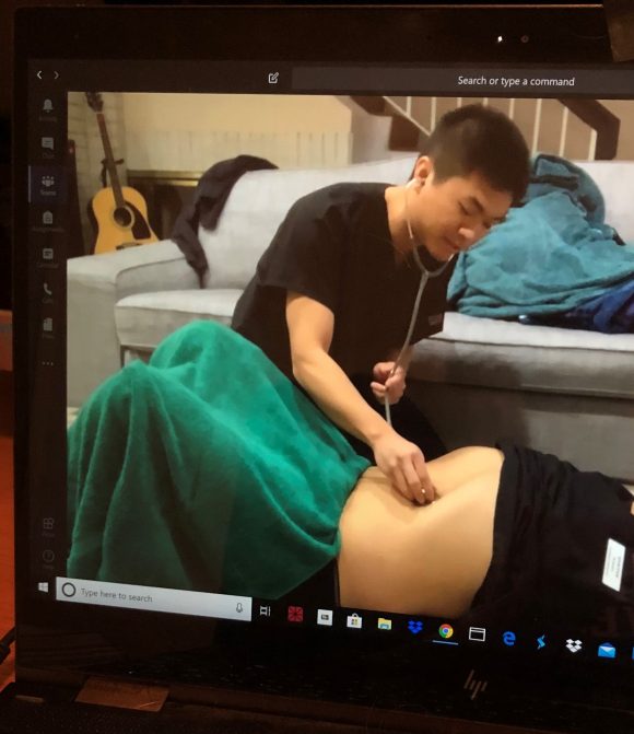 PA students who also happen to be roommates, practice abdominal exams while a professor watches via webcam. Typically, students wear gloves and masks. But given the demand for such protective gear in the healthcare community and since the students already share a household, they made do without.