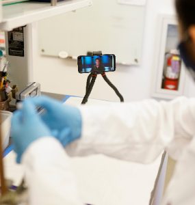 A chemistry student performs lab work as chemistry professor Chris Kim, Ph.D., provides mentorship and supervision through a mounted phone, tripod and body camera.