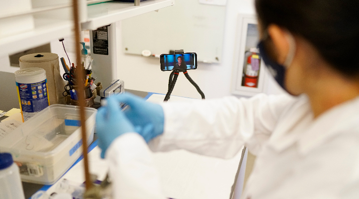 A chemistry student performs lab work as chemistry professor Chris Kim, Ph.D., provides mentorship and supervision through a mounted phone, tripod and body camera.