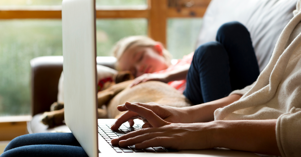 Woman working on a laptop while child sleeps next to her on the couch.