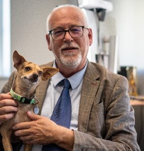 Photo of School of Comm Dean M. Andrew Moshier in university office with pet dog.