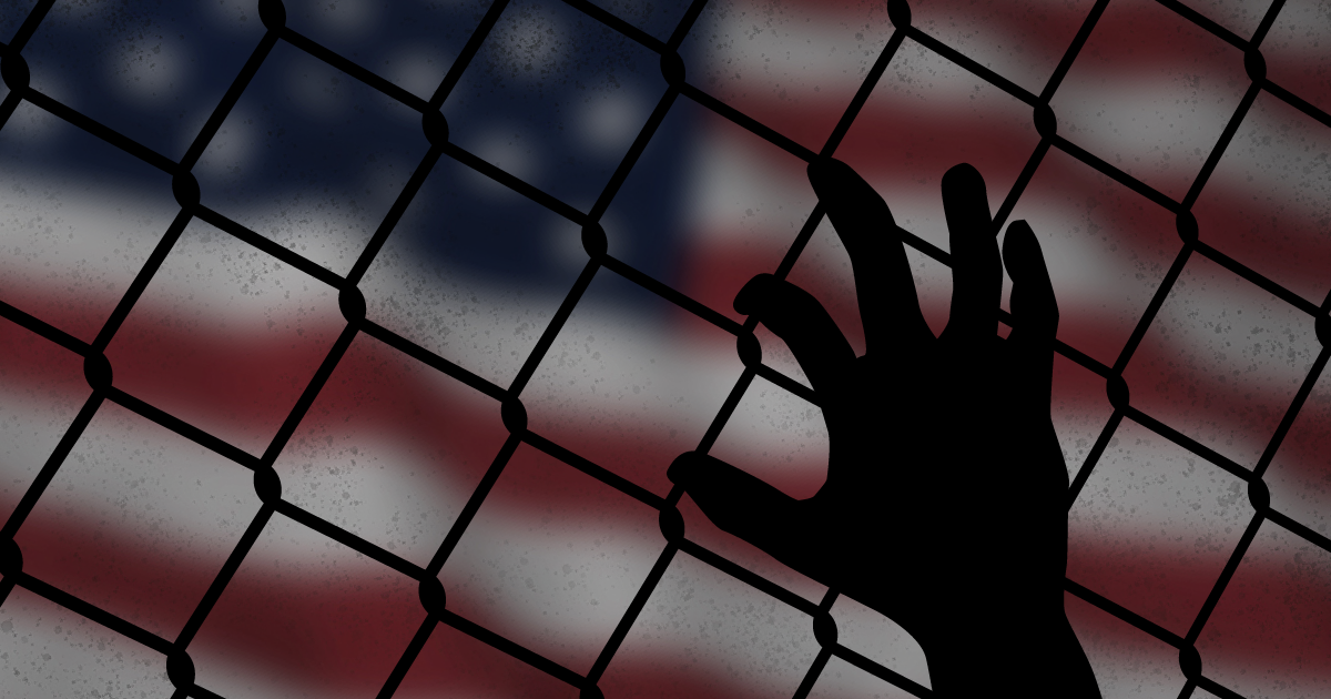a hand grasps at a chain link fence with the American flag in the background