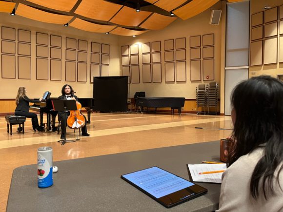 cellist sitting by a piano, in the foreground is the clinician with a notepad