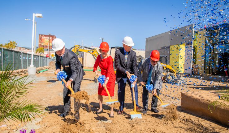 Four nicely dressed people in suits and hardhats digging into the ground of a construction site