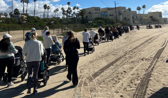 line of about 20 moms with strollers walking on beach