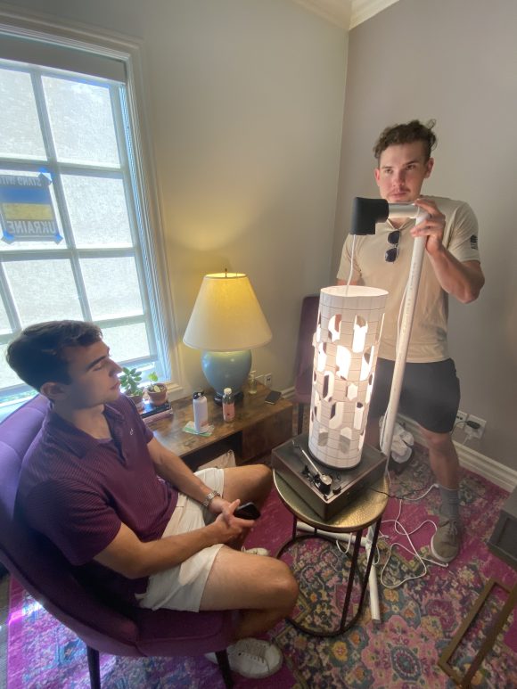 two male students in a living room with a device that looks like a paper cylinder attached to a record turntable, with a lightbulb suspended inside