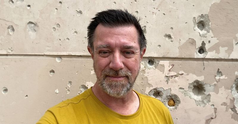 man wearing a yellow shirt standing against a wall with bullet holes.