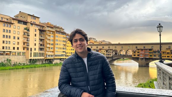 Summer Undergraduate Research Fellow Marcelo De La Maza Mendizabal stands on a balcony overlooking a river in Florence.