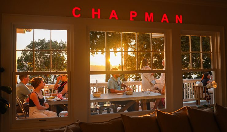 view out of a window into golden hour light, people on porch socializing, lights over window reading CHAPMAN