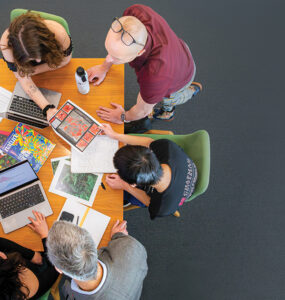 overhead shot of researchers gathered around table with copmuters, books and papers