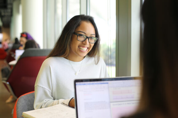 A young woman wearing glasses smiles while sitting at a table by a large window, with an open notebook and another student’s laptop visible in the foreground.