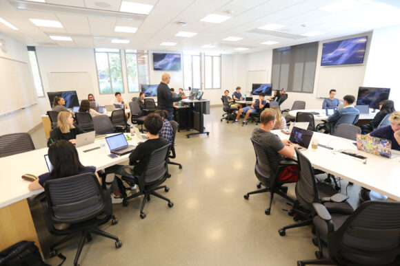 Students seated in groups at large tables use laptops while an instructor stands at the front of a bright, modern classroom with multiple screens and whiteboards.