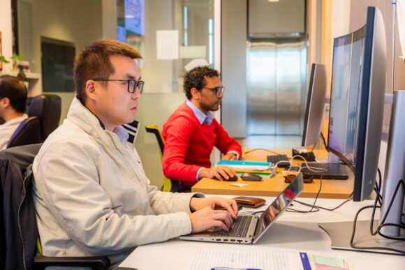 Two people sit at desks working intently on laptops and large monitors in a modern office-style lab environment.