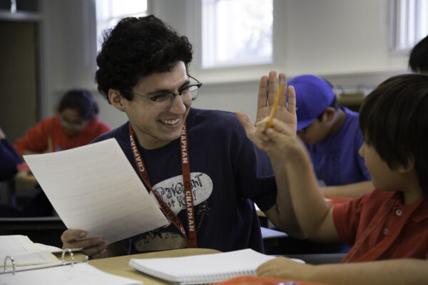 A college student smiles while assisting a child with schoolwork at a classroom desk.