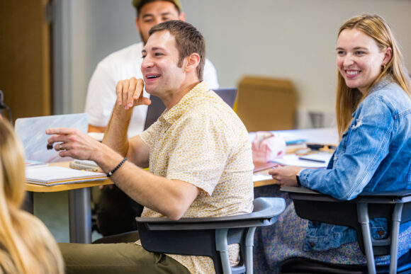 Adult students sit at desks, smiling and participating in a group discussion during class.