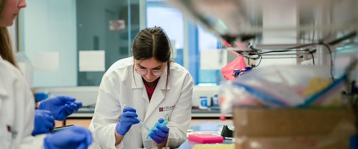 Two students in lab coats and gloves prepare samples at a laboratory bench using pipettes and scientific equipment.