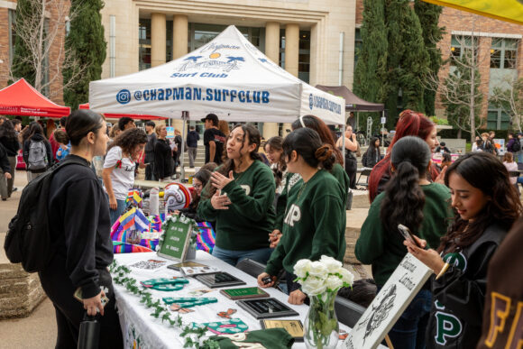 Outdoor campus event with a “Chapman Surf Club” canopy over an information table displaying flyers, stickers, and items for sign-ups, with students gathered around and other booths in the background.