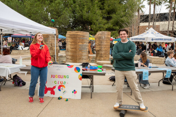 At an outdoor club fair, a student juggles colorful balls beside a hand-painted “Juggling Club” sign, while another student stands on a balance board near tables and tents.
