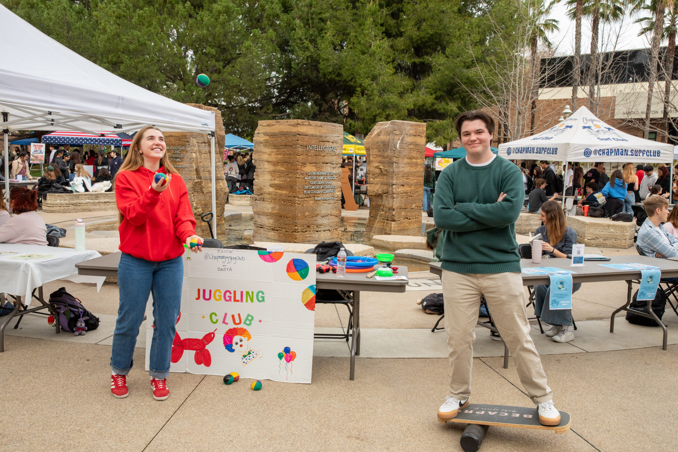 At an outdoor club fair, a student juggles colorful balls beside a hand-painted “Juggling Club” sign, while another student stands on a balance board near tables and tents.