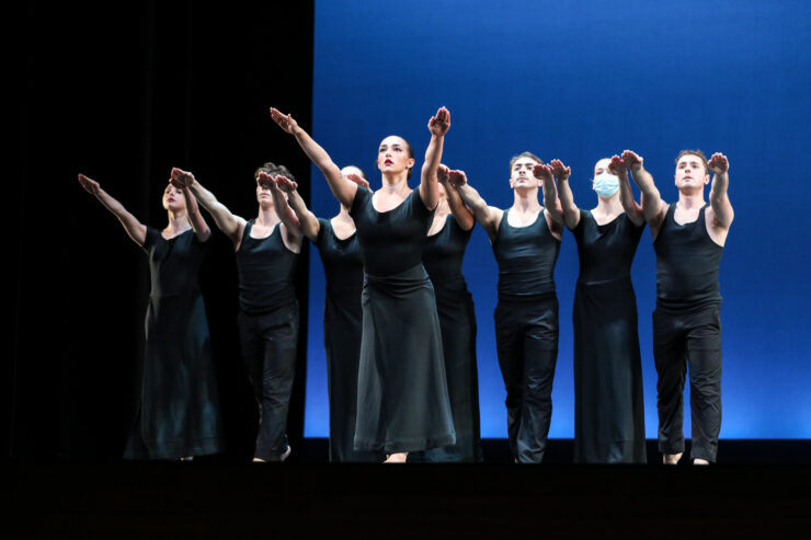 A group of dancers in black costumes perform onstage against a blue backdrop, standing in formation with their arms extended forward.