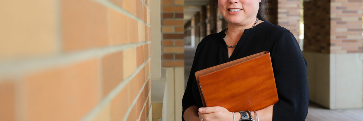 Stephanie Takaragawa, associate professor of sociology, stands next to a brick wall with folder in hand.