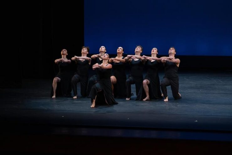 Student dancers in black costumes kneel in a tight formation onstage, arms linked and lifted, performing against a blue backdrop.