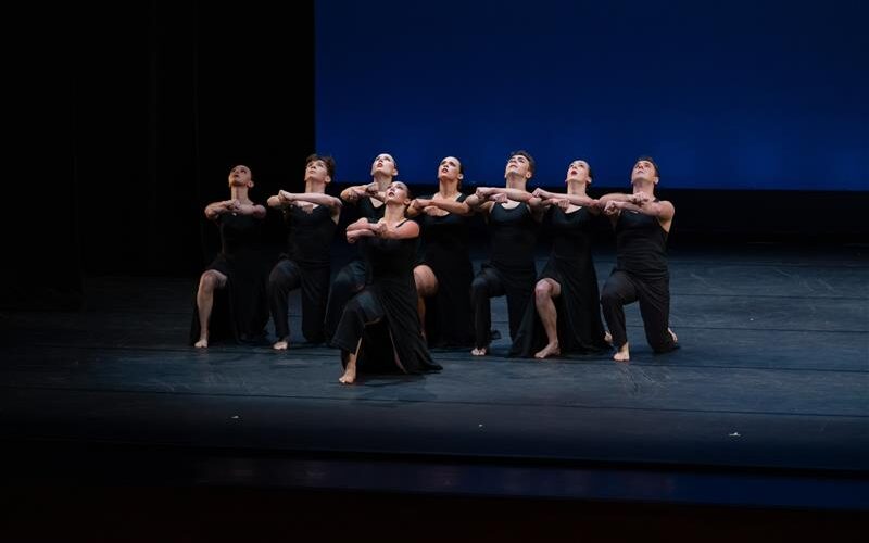 Student dancers in black costumes kneel in a tight formation onstage, arms linked and lifted, performing against a blue backdrop.