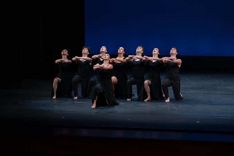 Student dancers in black costumes kneel in a tight formation onstage, arms linked and lifted, performing against a blue backdrop.