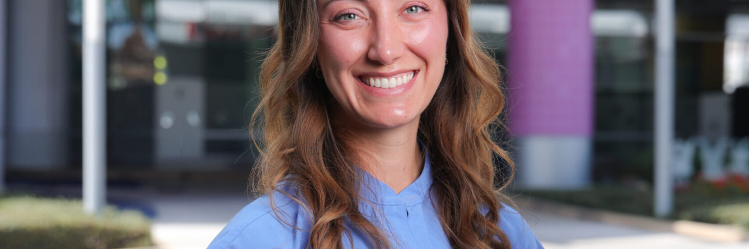 Erika Ebe stands outdoors in blue medical scrubs in front of the CHOC hospital sign.