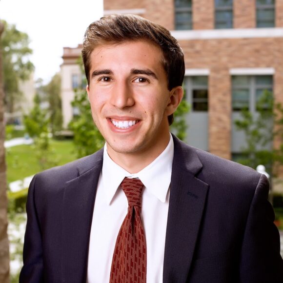 Adam Kalawi smiling in a dark suit and red tie outdoors with a campus building in the background.