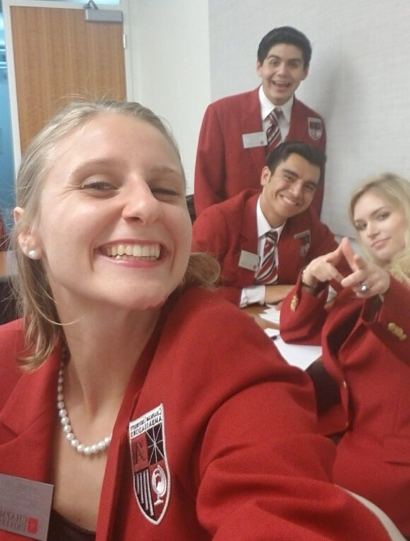 A group selfie taken indoors showing several people wearing matching red blazers with striped ties, seated at a table.