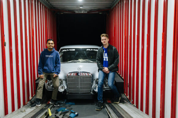 Two people stand in a red-and-white enclosed trailer in front of a classic Mercedes with “AMG” lettering on the windshield.