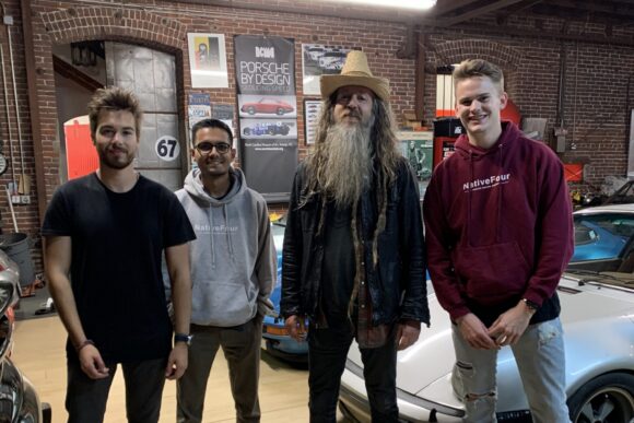 Four people stand in a garage beside sports cars and automotive posters, including a person with a long beard and a hat.