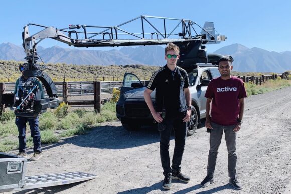  Two people stand on a dirt road beside a vehicle fitted with a large camera crane rig, with mountains in the background under a clear sky.
