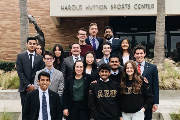 A group of people dressed in business attire and Alpha Kappa Psi clothing pose on steps outside the Harold Hutton Sports Center on Chapman’s campus.