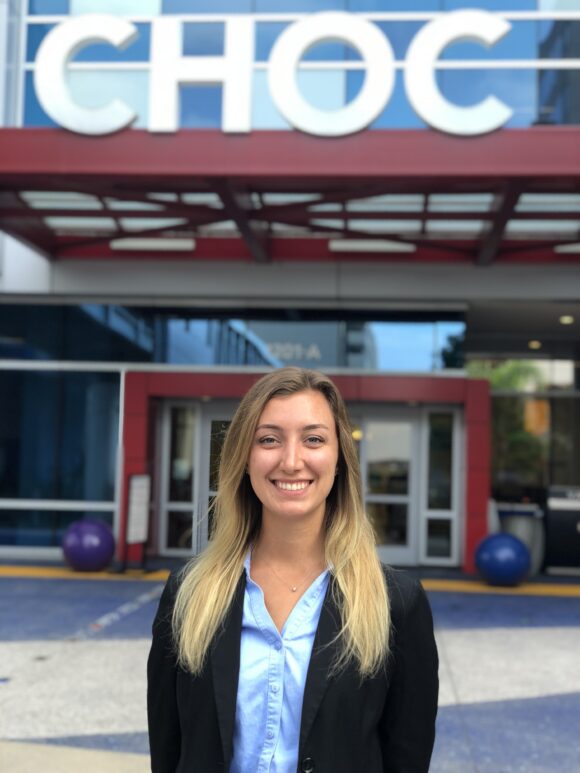 Erika Ebe stands in front of the CHOC hospital entrance sign, wearing a black blazer and light blue shirt.