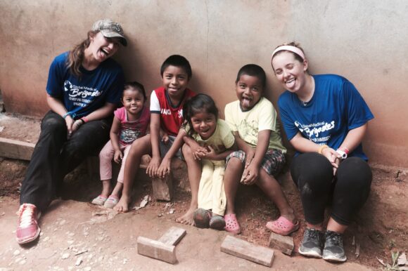 Erika Ebe and another volunteer in blue Global Medical Brigades shirts sit on a bench with several children against a concrete wall outdoors.