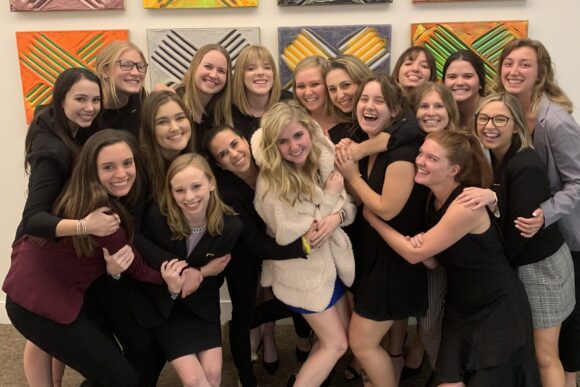 A large group of students pose closely together indoors at a formal event, with colorful square artwork displayed on the wall behind them.