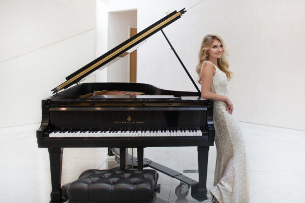 A person in a light-colored formal dress stands beside a grand piano with its lid open in a bright indoor space.