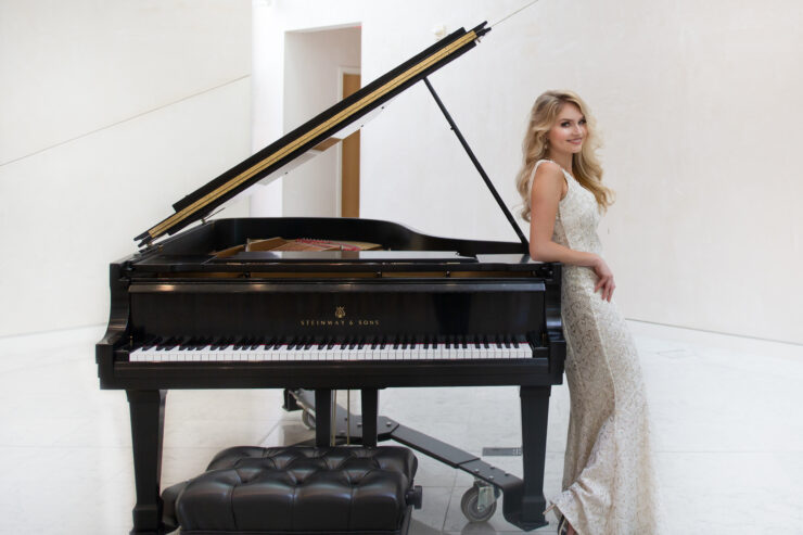 A person in a light-colored formal dress stands beside a grand piano with its lid open in a bright indoor space.