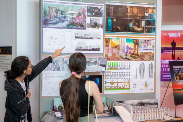 Two people stand in an indoor exhibit space looking at a large display board of architectural renderings, floor plans, and diagrams; one person points to a drawing while the other, with a long ponytail, watches.
