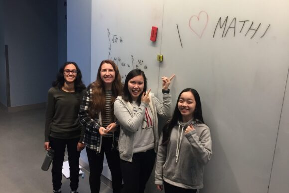 Arshia Sharma stands with three classmates near a whiteboard that reads “I heart math!” in a classroom.
