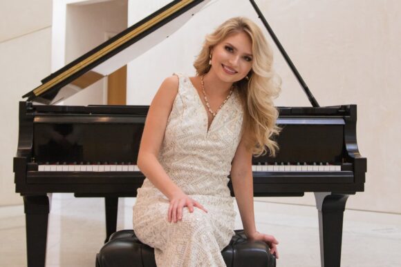 A person in a light-colored formal dress sits on a piano bench in front of a grand piano in a bright indoor space.