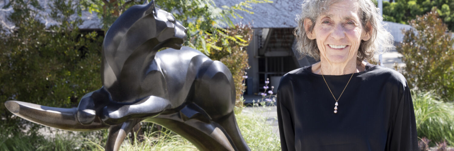 Janeen Hill stands smiling outdoors next to a bronze panther sculpture, wearing a black long-sleeve top, with trees and a building visible in the background.