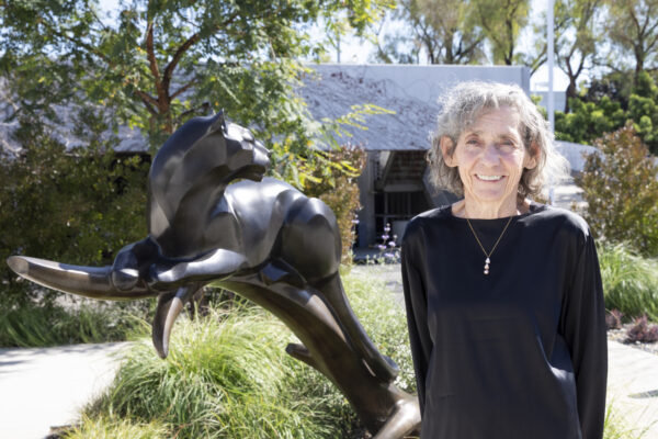 Janeen Hill stands smiling outdoors next to a bronze panther sculpture, wearing a black long-sleeve top, with trees and a building visible in the background.