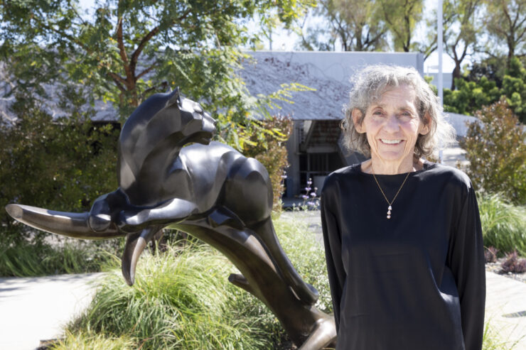 Janeen Hill stands smiling outdoors next to a bronze panther sculpture, wearing a black long-sleeve top, with trees and a building visible in the background.