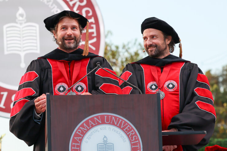 Ross Duffer, left, and his twin brother, Matt Duffer, stand together at a Chapman University podium in academic regalia, sharing a laugh during their keynote address.