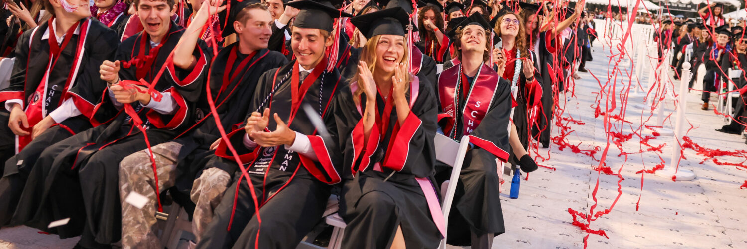 Alt text: Rows of Chapman University graduates in caps and gowns cheer and laugh as red streamers and confetti rain down on them during the Commencement ceremony at Wilson Field, with a packed stadium visible in the background.