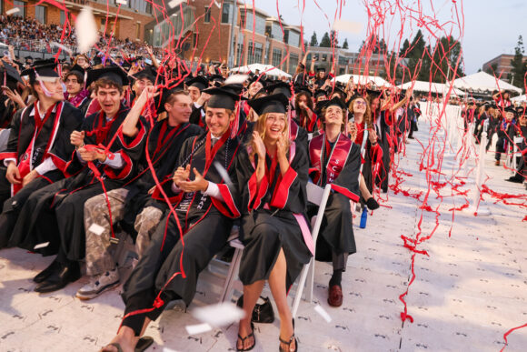 Alt text: Rows of Chapman University graduates in caps and gowns cheer and laugh as red streamers and confetti rain down on them during the Commencement ceremony at Wilson Field, with a packed stadium visible in the background.